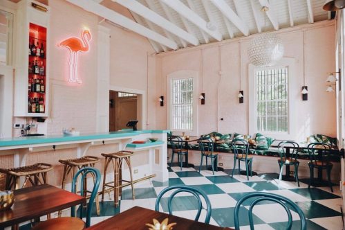 Bright, stylish café interior with pink walls, a neon flamingo sign, and checkered flooring. Cozy seating area visible.