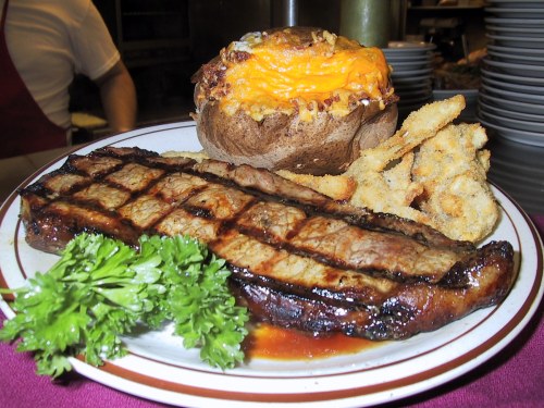 A grilled steak with a baked potato topped with cheese and fried okra, garnished with parsley on a plate.