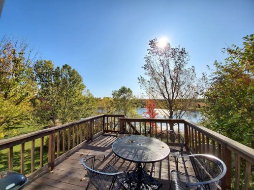 A sunny deck with a table and chairs overlooks a serene lake surrounded by colorful autumn trees.
