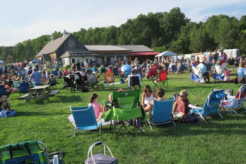 A large crowd gathers on a grassy field with chairs, enjoying an outdoor event near a rustic building.