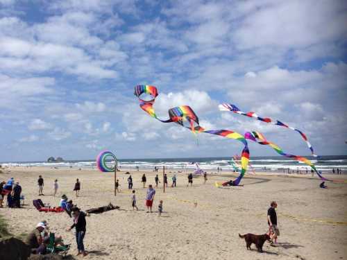 Colorful kites fly over a busy beach with people enjoying the sun and waves on a partly cloudy day.