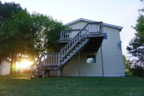 A two-story house with a wooden staircase, surrounded by trees, and sunlight shining in the background.