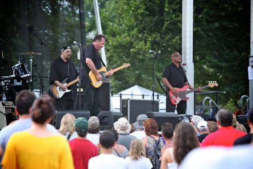 A band performs on stage in front of an audience at an outdoor event, with trees in the background.