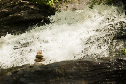 A small stack of stones sits on a rock in front of a rushing waterfall, surrounded by lush greenery.