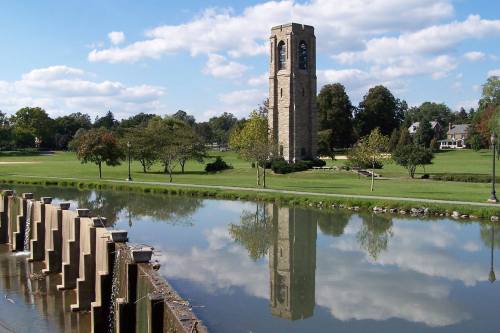 A stone tower beside a calm lake, surrounded by trees and a grassy park under a blue sky with fluffy clouds.