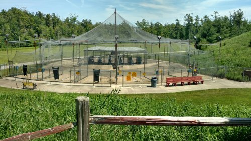 A large fenced enclosure with a netted roof, surrounded by grass and trees, featuring benches and a trash can nearby.
