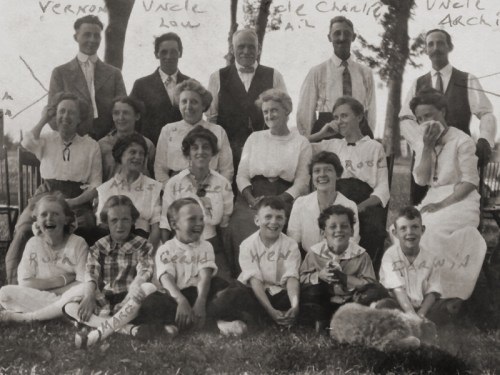 A vintage family photo featuring adults and children, posed outdoors with trees in the background. Names are handwritten on the image.