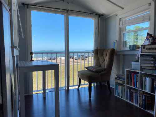 Cozy interior with a chair and bookshelf, featuring a large window overlooking a beach and blue sky.