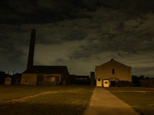 A dark, cloudy night scene featuring two large buildings and a tall chimney, with a grassy area in the foreground.