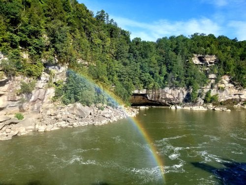 A vibrant rainbow arcs over a river, surrounded by lush green cliffs and trees under a clear blue sky.