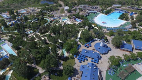 Aerial view of a water park featuring slides, a wave pool, and lush greenery with blue umbrellas scattered around.