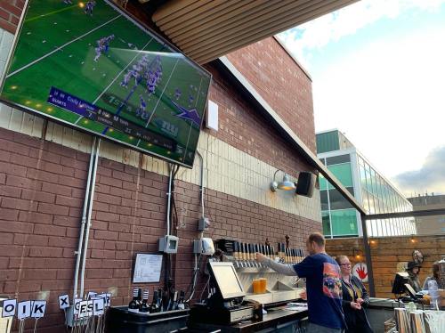 A bar with a large TV showing a football game, and a bartender serving drinks to customers outdoors.
