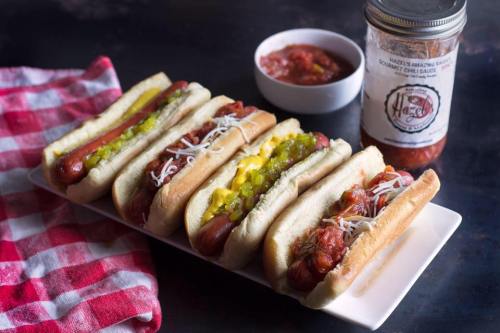 A platter of five hot dogs topped with various condiments, alongside a jar of sauce and a small bowl of salsa.