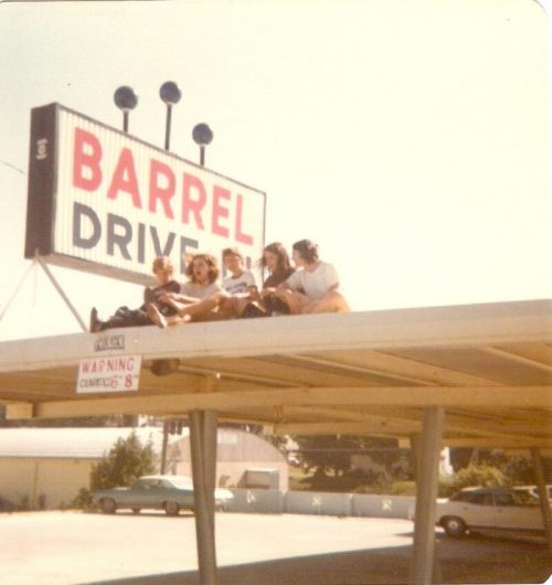 A group of people sitting on a roof near a "Barrel Drive" sign, with cars parked below in a sunny setting.