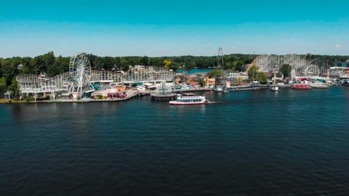 Aerial view of a vibrant amusement park by the water, featuring rides, boats, and lush greenery in the background.