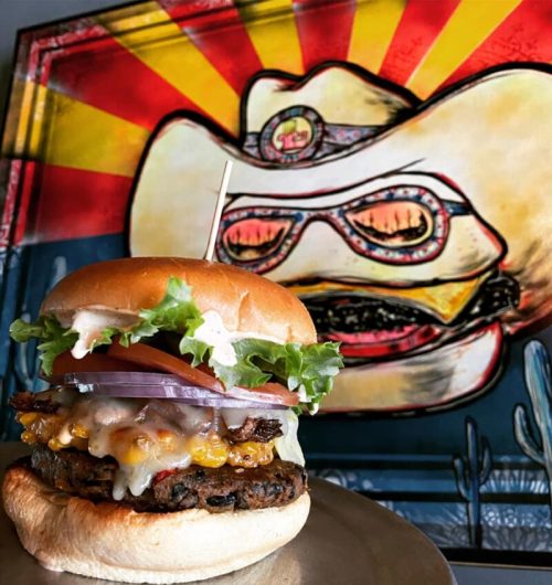 A delicious burger with lettuce, tomato, onion, and melted cheese, set against a colorful cowboy-themed mural.