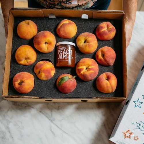A box of fresh peaches surrounding a jar of peach jam on a marble surface.