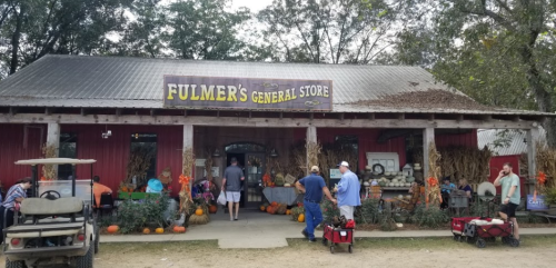 A rustic general store with a sign reading "Fulmer's General Store," surrounded by pumpkins and visitors.