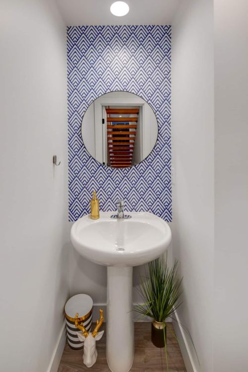 A modern bathroom with a round mirror, patterned blue wall, sink, and decorative plant.