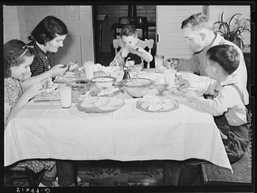 A family of five sits around a table, enjoying a meal together, with plates of food and drinks in front of them.