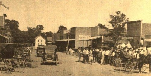 Historic street scene with early automobiles, horse-drawn carriages, and a crowd gathered in a small town.