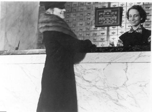 A woman in a fur coat smiles at a hotel receptionist behind a marble counter.