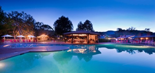 A serene pool area at dusk, featuring a gazebo, lounge chairs, and surrounding trees, reflecting in the water.