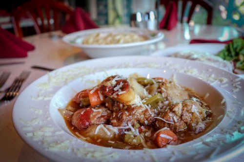 A plate of hearty stew with vegetables and meat, topped with cheese, set on a dining table with pasta in the background.