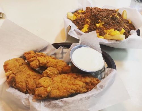 A plate of crispy fried chicken tenders with a side of ranch dressing, next to a dish of loaded cheese fries.