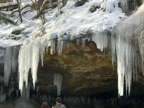 Two people stand beneath a rocky overhang adorned with icicles and snow, surrounded by a winter landscape.