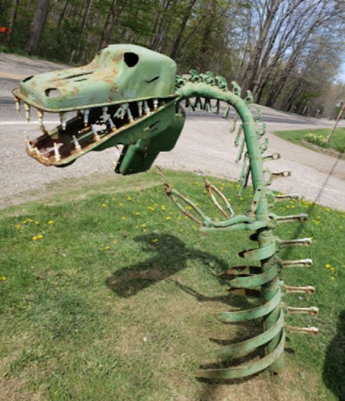 A whimsical dinosaur sculpture made from green metal, featuring a long spine and a large head, set in a grassy area.