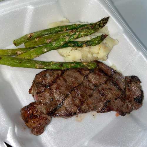 A grilled steak with grill marks, served with mashed potatoes and asparagus on a white takeout container.