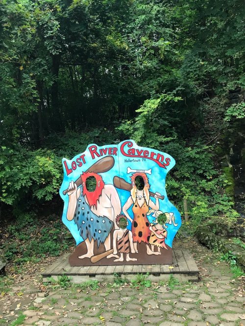 Colorful sign for Lost River Caverns featuring cartoon cavemen in a forested area.