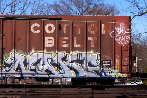 A rusted train car with "COTTON BELT" painted on it, covered in colorful graffiti. Trees are visible in the background.