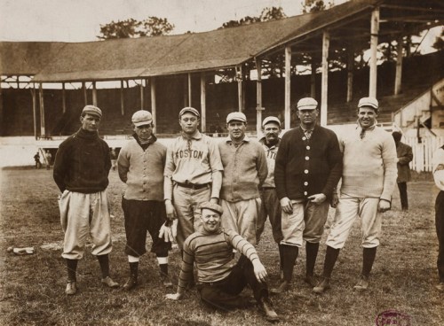 A historic baseball team poses on the field, wearing vintage uniforms and caps, with a stadium in the background.