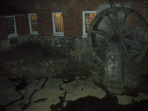 A dark image of a stone structure with a large water wheel beside it, illuminated by nearby windows.