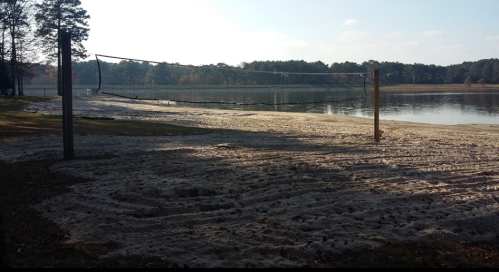A sandy beach area with a volleyball net near a calm lake, surrounded by trees and a clear sky.