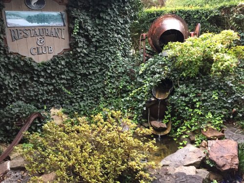 A rustic restaurant sign surrounded by lush greenery and a water feature with cascading pots.