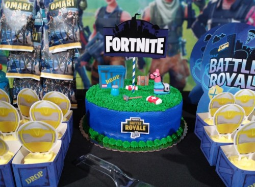 A colorful Fortnite-themed cake with green frosting, decorations, and themed treats on a table.