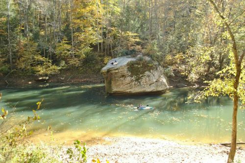 A serene river scene with a large rock, surrounded by trees, and a person kayaking in the water.