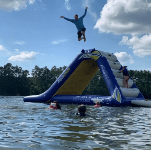 A person jumps off an inflatable water slide into a lake, while others swim nearby on a sunny day.
