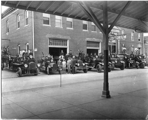 Historic photo of a group of people and vintage fire trucks in front of a brick auditorium building.
