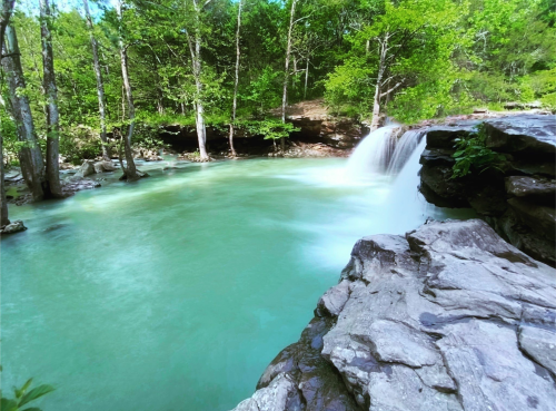 A serene waterfall cascades into a turquoise pool, surrounded by lush green trees and rocky terrain.