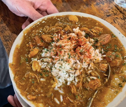 A plate of gumbo with shrimp, sausage, and rice, garnished with herbs and spices, served on a wooden table.
