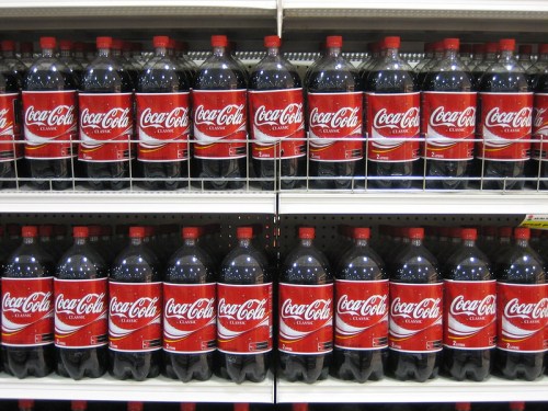 Rows of large Coca-Cola bottles on a store shelf, showcasing the classic red label design.