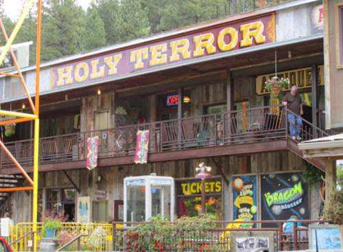 A rustic building with "Holy Terror" sign, featuring antique shops and a balcony, surrounded by trees.
