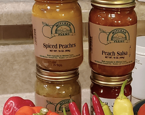 Three jars of Mitcham Farms products: Spiced Peaches, Peach Salsa, and a third jar, with colorful peppers in the foreground.