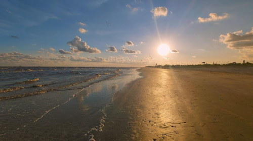 A serene beach at sunset, with golden sand reflecting the sun and gentle waves under a blue sky with scattered clouds.