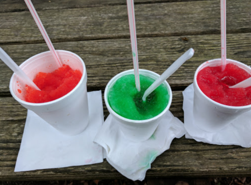 Three cups of colorful slushies—red, green, and red—on a wooden table with straws and napkins.
