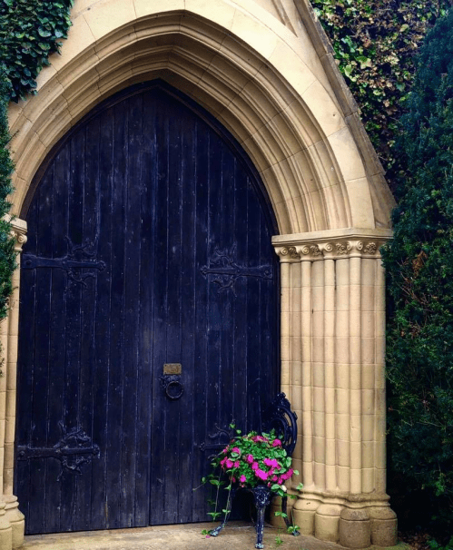 A large, arched wooden door framed by stone columns and ivy, with a decorative bench holding pink flowers nearby.